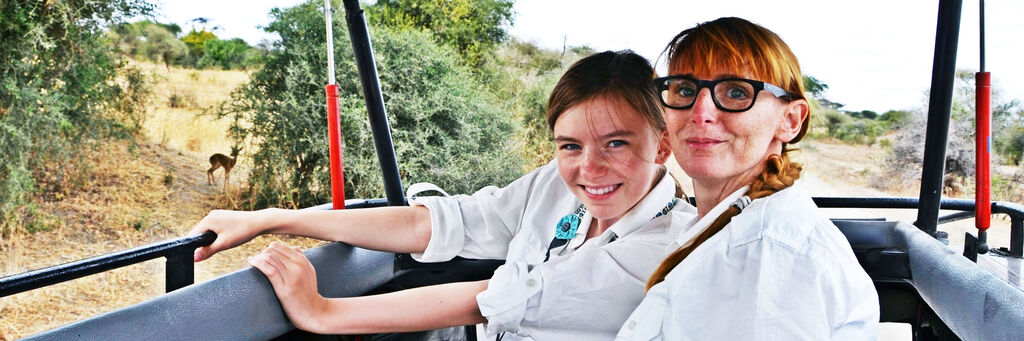 Nicole Hildebrandt und ihre Tochter Luzia lächeln im Safari-Jeep gemeinsam in die Kamera. Links im Hintergrund eine Gazelle sowie Buschwerk.