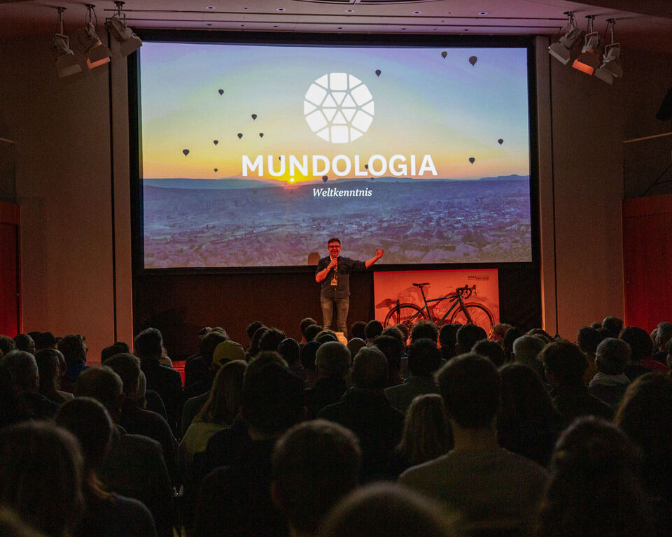 Ein Mann steht auf einer Bühne. Hinter ihm ist eine Leinwand, auf dem das Mundologia-Logo zu sehen ist.