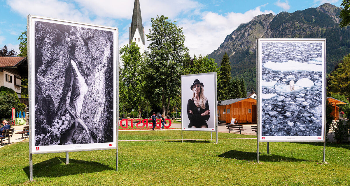 Auf einer Wiese stehen drei große Fotografien. Im Hintergrund ist eine Kirche zu sehen.