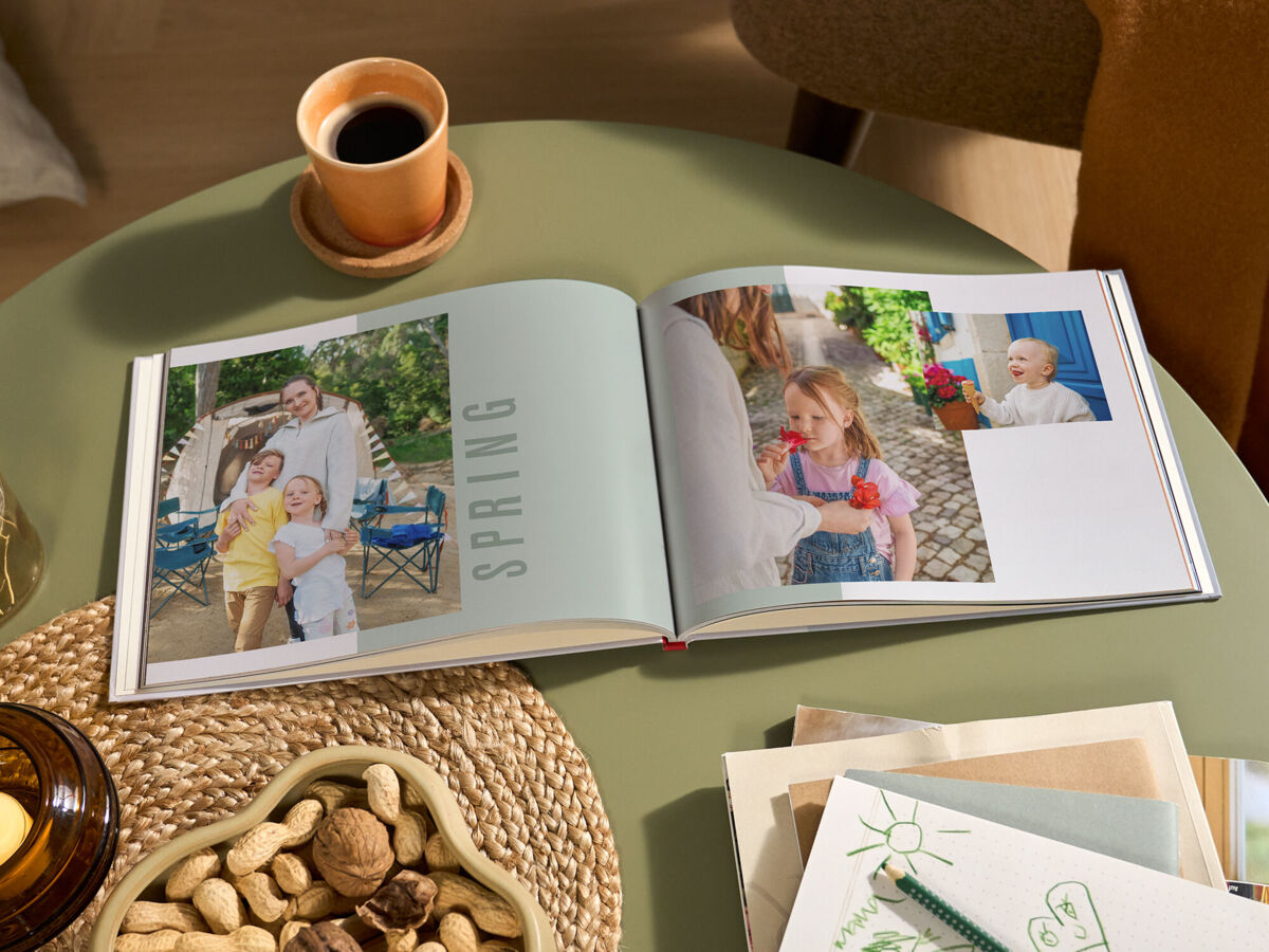 Aufgeschlagenes CEWE FOTOBUCH mit Bildern einer Familie und der Aufschrift „SPRING“ auf einem grünen Tisch; daneben stehen eine Tasse Kaffee