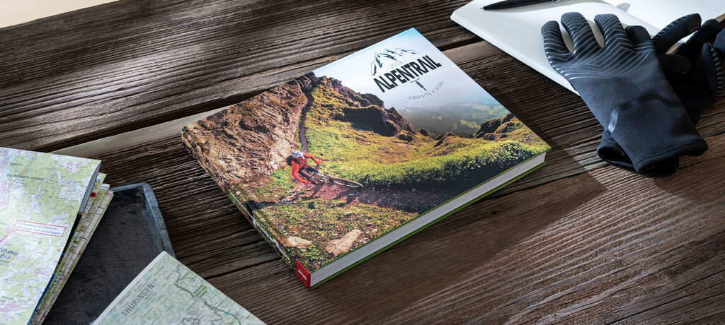 Ein CEWE FOTOBUCH liegt auf einem Holztisch, umgeben von Karten und Handschuhen. Es zeigt eine Berglandschaft mit einem Radfahrer.
