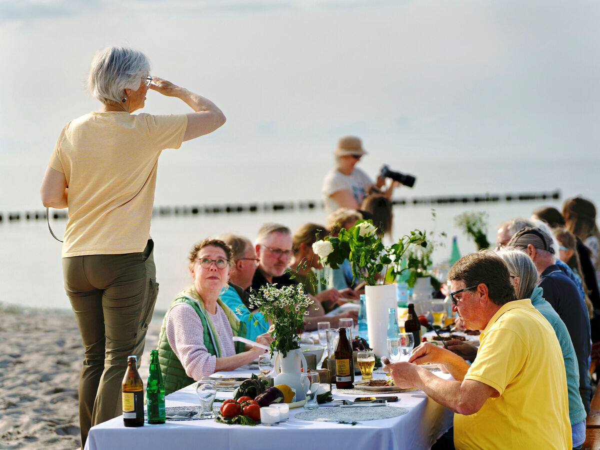 An einem Strand ist ein langer Tisch aufgebaut, an dem mehrere Menschen sitzen und gemeinsam essen und trinken.