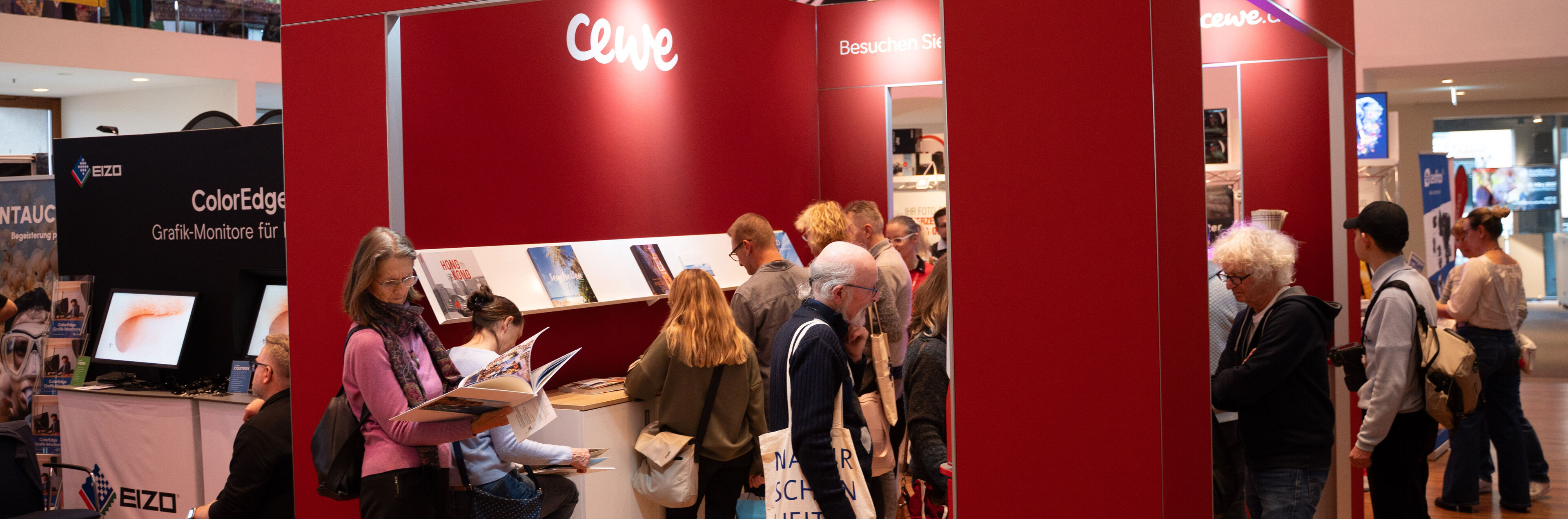 In einer Halle ist ein Messestand von CEWE aufgebaut. Viele Leute betrachten den Stand und die ausgelegten Fotobücher.