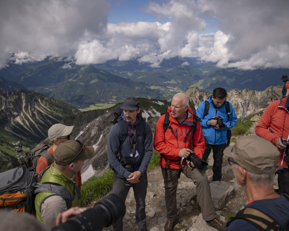 Mehrere Personen in Outdoor-Outfits und mit Kameras in den Händen stehen diskutierend auf einem Berggipfel.