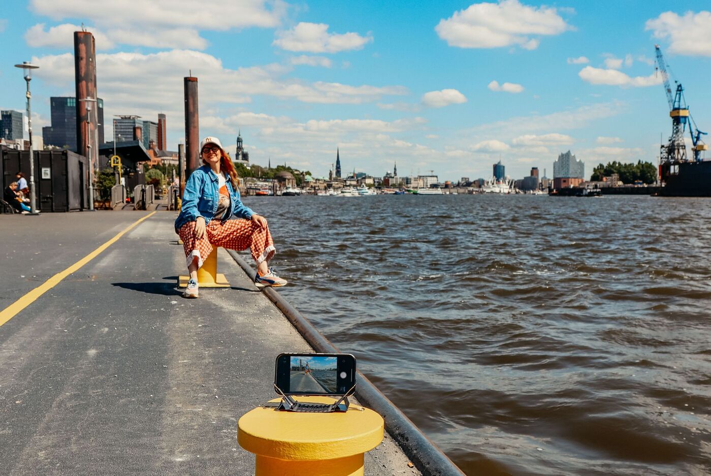 Person am Wasser sitzt auf einem Poller, sonniger Tag, Hamburg im Hintergrund, Handy mit Stativ vorne