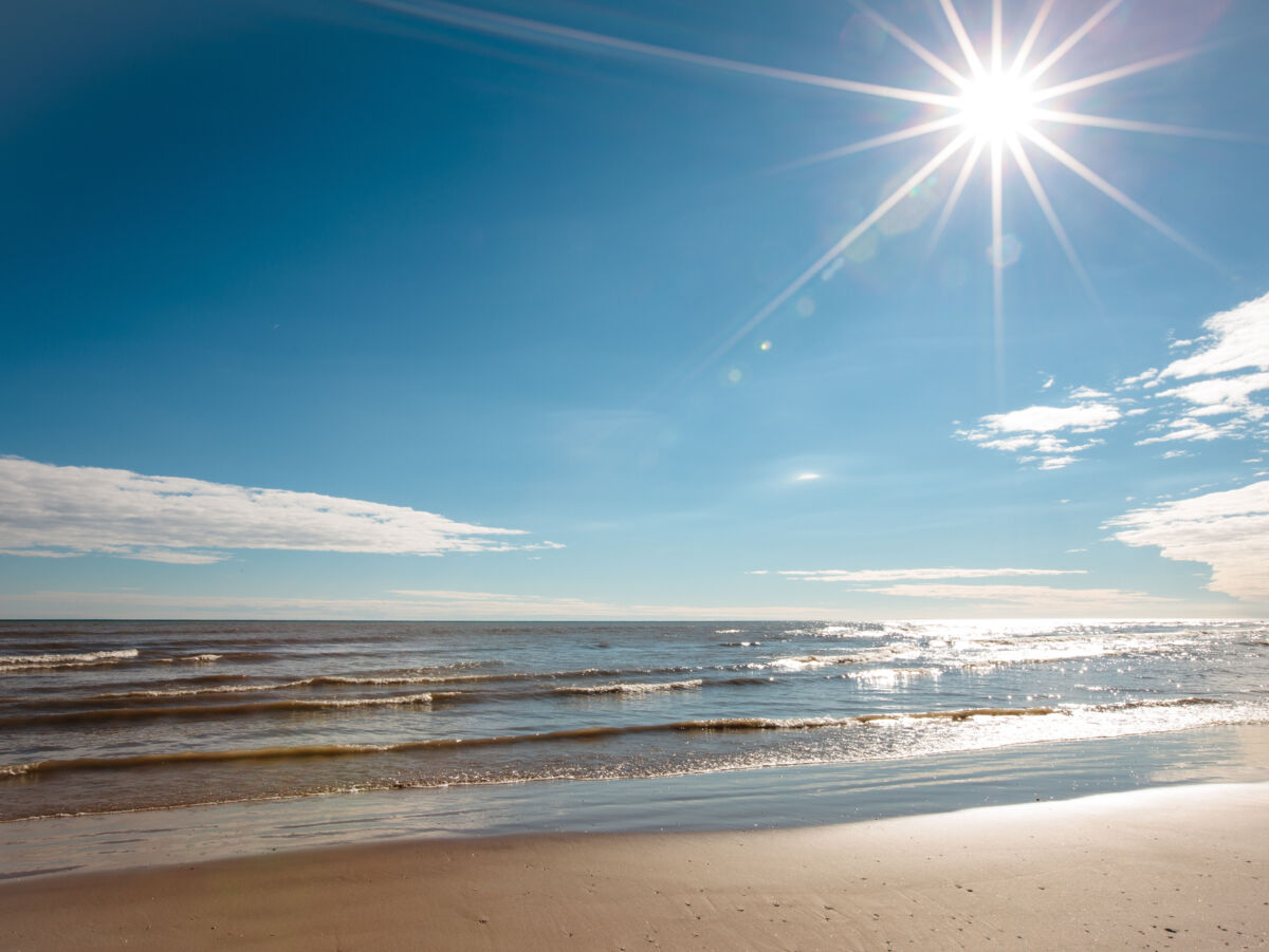 Sonnenstern am blauen Himmel über Meer, das in sanften Wellen an den Strand spült