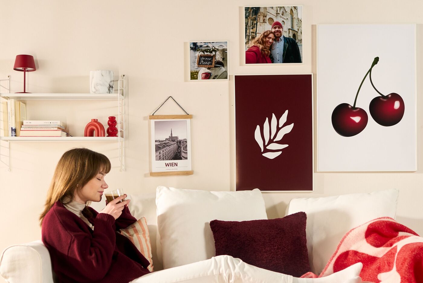 Eine Frau sitzt auf dem Sofa mit einem Getränk, rote Bilder und Poster hängen an der Wand.