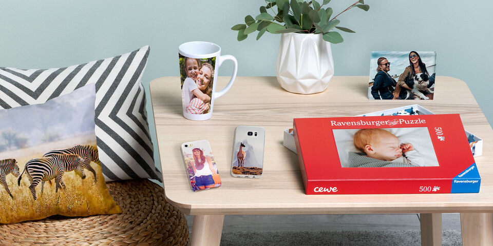 Ein Tisch mit verschiedenen Fotoprodukten: Ein CEWE Fotobuch, eine Tasse mit einem Foto, Handyhüllen und ein Puzzle. Ein Kissen mit Zebramuster und Pflanzen im Hintergrund ergänzen die Szene.