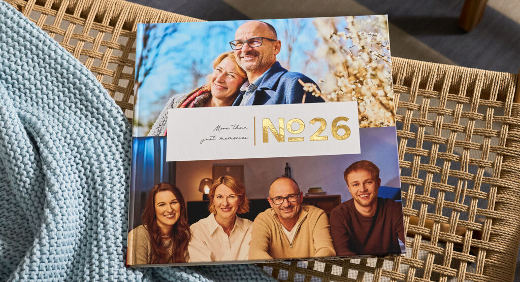 CEWE FOTOBUCH „N°26“ mit Fotos eines Paares und einer Familie von vier Personen auf einem geflochtenen Stuhl mit blauer Decke.