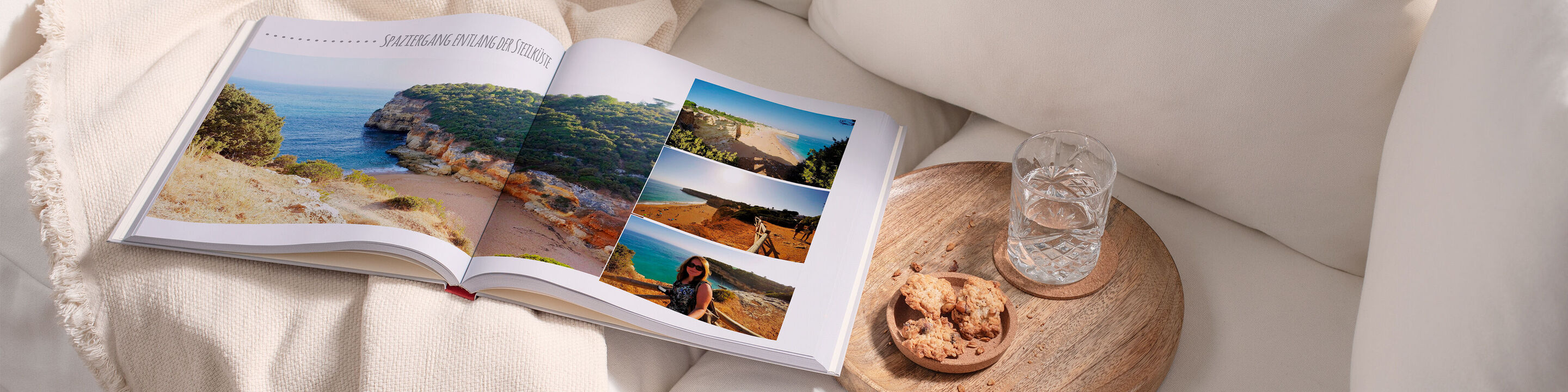 Ein offenes Fotobuch mit Urlaubsfotos auf einem Tisch mit Keksen und einem Glas Wasser. Der Tisch ist mit einer weichen Decke verziert.