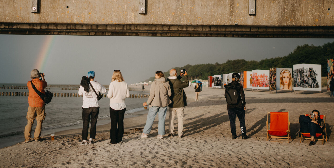 Fotografinnen und Fotografen stehen am Strand und fotografieren einen Regenbogen über der Ostsee. Im Hintergrund ist eine Fotoausstellung.