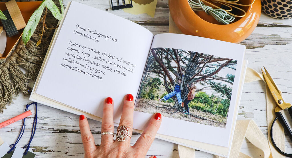 Eine Hand mit roten Nägeln blättert durch ein offenes Fotobuch, auf einer Holzoberfläche liegen Bastelmaterialien.