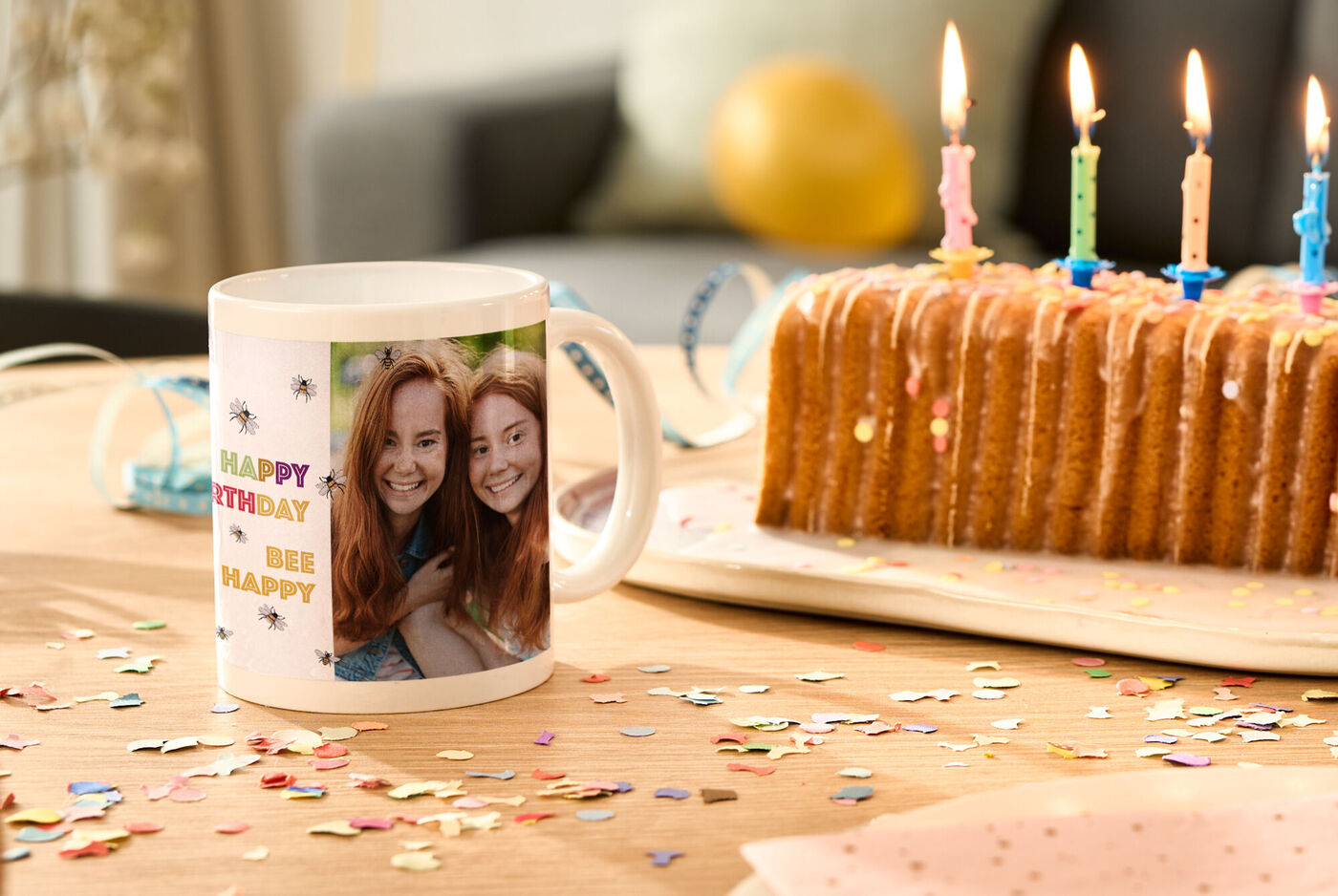 Eine Tasse mit einem bunten Geburtstagsdesign steht neben einer Geburtstagstorte mit Kerzen auf einem Tisch. Der Tisch ist mit buntem Konfetti dekoriert.