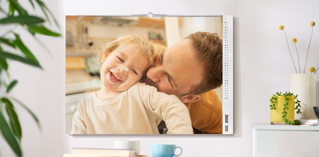CEWE Fotokalender mit einem Familienfoto hängt an einer hellen Wand über einem Tisch mit Tasse und Dekoration