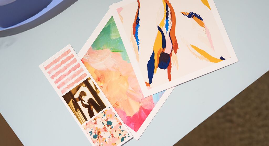 Mehrere bunte CEWE Fotodrucke mit abstrakten Designs und Mustern liegen auf einem hellblauen Tisch
