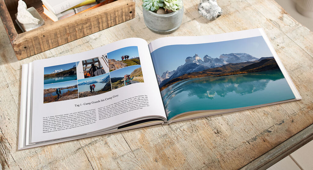 Offenes CEWE FOTOBUCH auf einem Holztisch mit Reisebildern von den Bergen und einem klaren See.