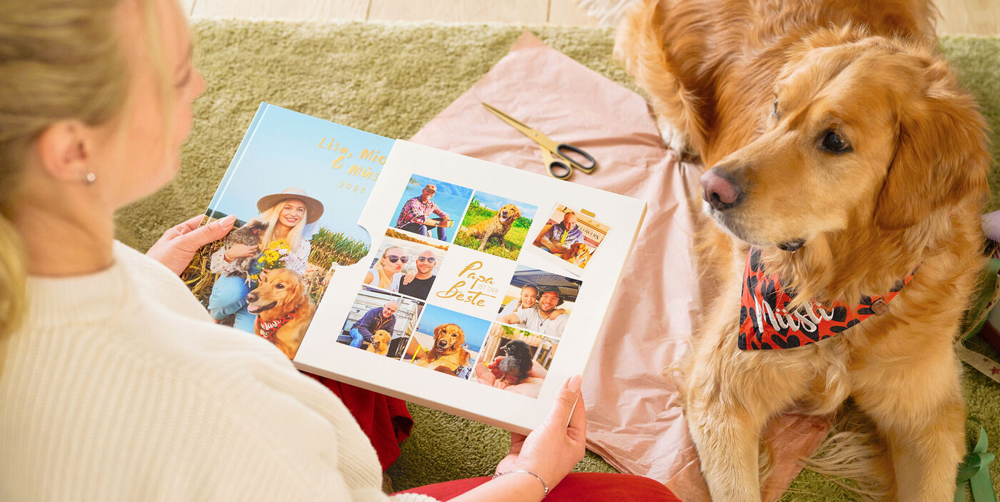Eine Frau hält ein CEWE FOTOBUCH mit Familienfotos in den Händen. Vor ihr liegt ein Hund und schaut sie an.