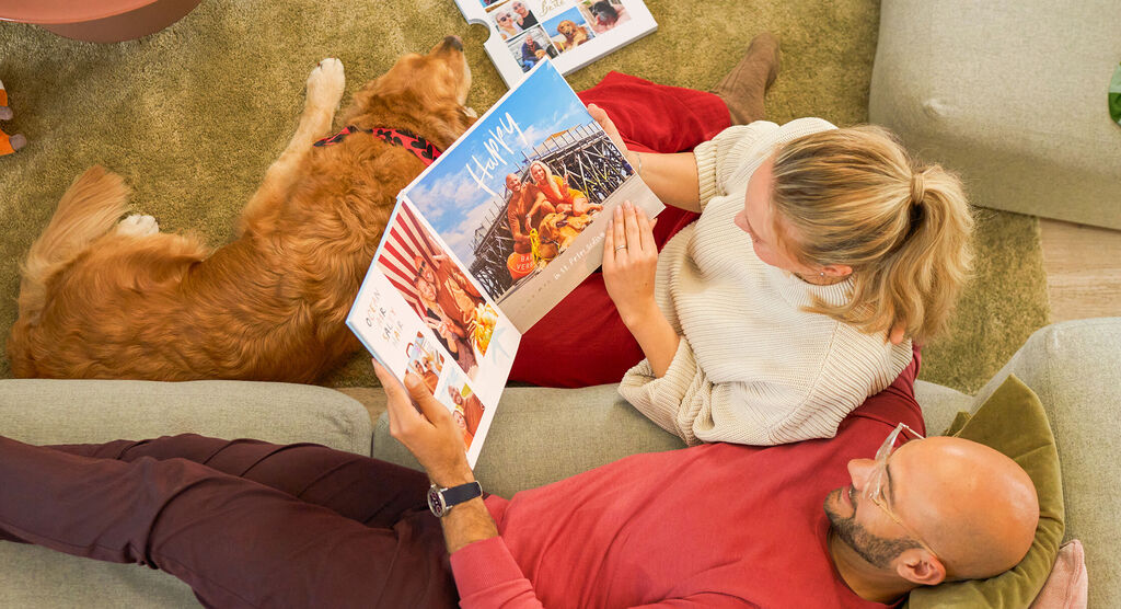 Eine Person liegt auf dem Sofa, eine andere sitzt davor. Sie blättern durch ein CEWE FOTOBUCH, ein Hund liegt daneben.