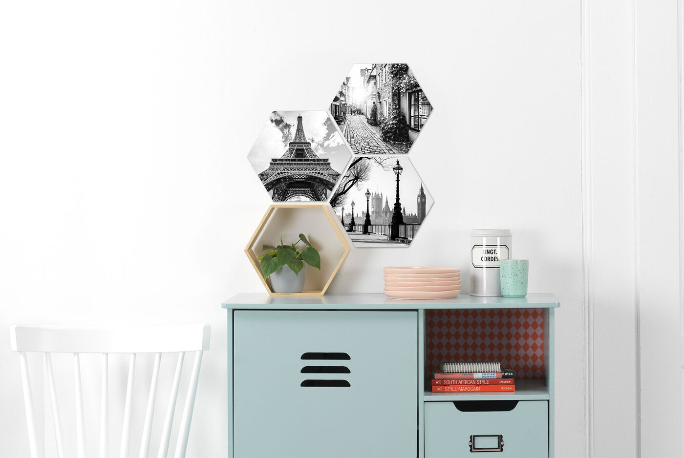 Verschiedene hexagonale Wandbilder mit schwarz-weißen Motiven hängen an einer weißen Wand über einem hellblauen Sideboard, auf dem Pflanzen und Dekoration stehen.