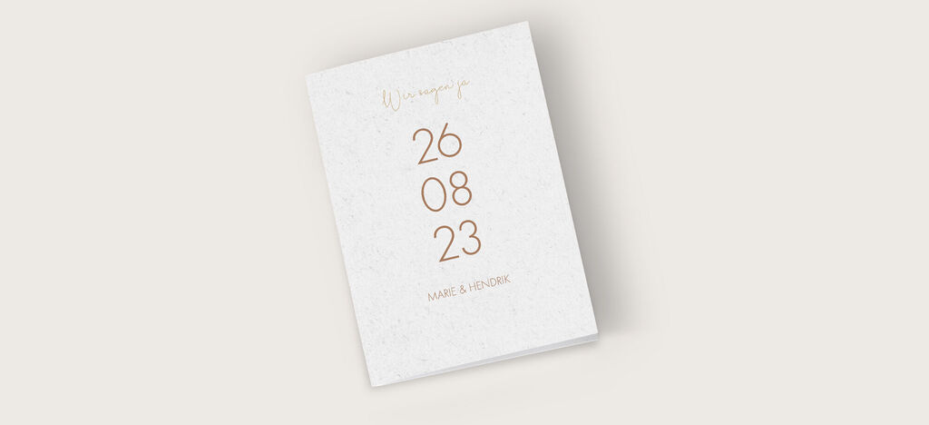 Ein elegantes weißes Fotobuch mit goldenen Akzenten und den Namen "Marie & Hendrik" sowie dem Datum "26.08.23" embossed auf dem Cover