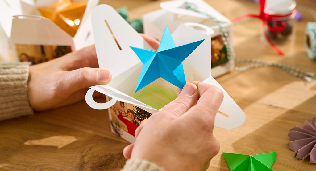 Zwei Hände stecken einen bunten Papierstern in eine Geschenkbox mit Fotomotiv