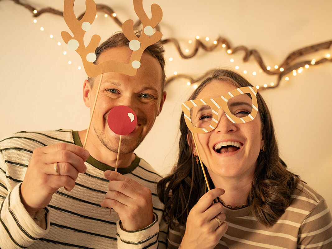 Ein fröhliches Paar hält Pappmasken vor dem Gesicht. Sie tragen Weihnachtsdeko mit Rentierohren. Im Hintergrund strahlen Lichter.
