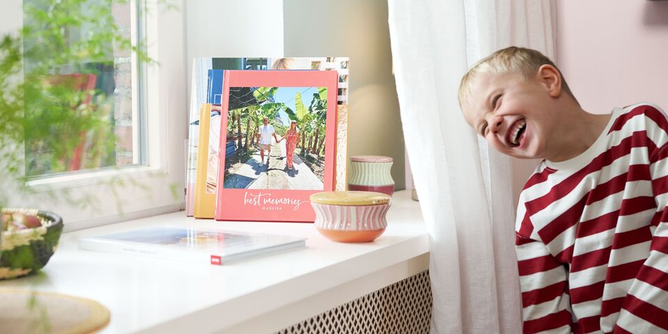 Ein lachernder Junge steht neben einer Fensterbank in einem hellen Wohnraum. Vor dem Fenster stehen einige farbige Fotobücher.