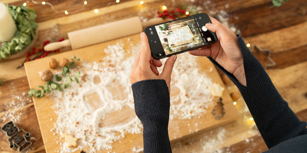Eine Person fotografiert mit einem Smartphone eine Auslage mit Mehl und Keksausstechern auf einem Holzbrett, um eine festliche Atmosphäre zu schaffen. Die Szenerie ist liebevoll gestaltet.