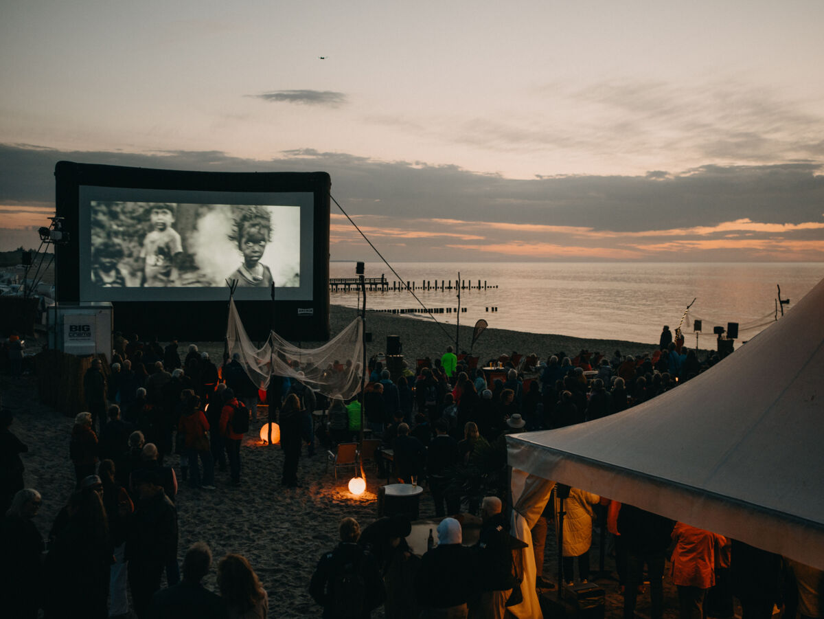 Auf einer Leinwand am Strand werden Fotos einer Ausstellung gezeigt. Vor der Leinwand sitzen Menschen im Sand.