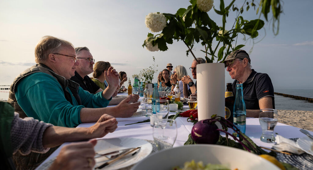 Dieses Foto zeigt den langen Party-Tisch am Strand. Es sind Menschen zu sehen, die essen und sich dabei unterhalten.