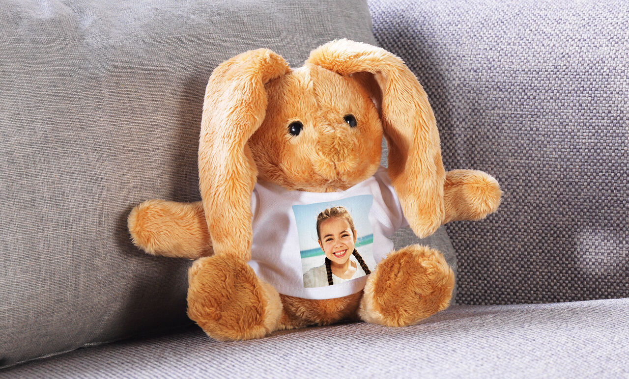 Ein süßes, plüschiges Kaninchen sitzt auf einer Couch. Es trägt ein T-Shirt mit einem Foto darauf.
