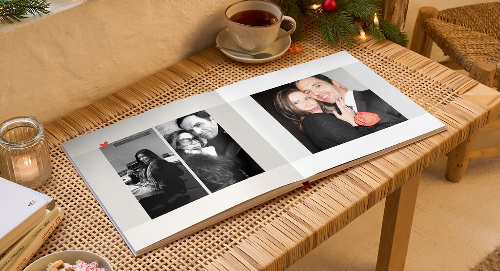 Ein offen liegendes Fotobuch auf einem Tisch. Es zeigt zwei Fotos von glücklichen Menschen. Auf der linken Seite ist ein Paar zu sehen, auf der rechten Seite sind zwei Frauen, die lächeln. Eine bequeme Tasse steht auf dem Tisch. Im Hintergrund sind Weih