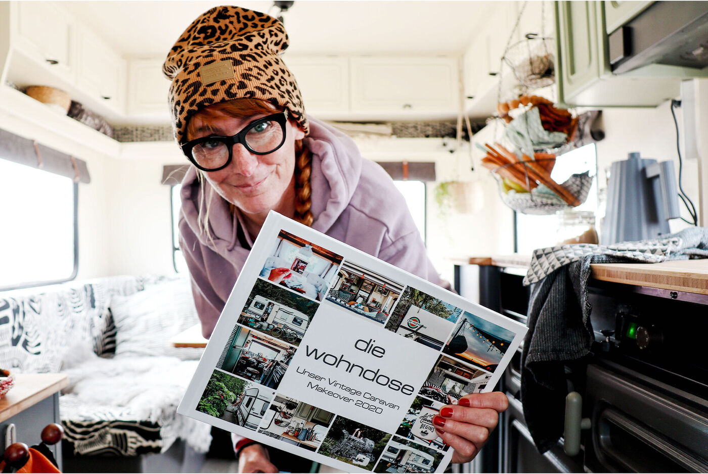Frau hält ein CEWE Fotobuch mit dem Titel 'Die Wohndose' in der Hand. Im Hintergrund ist eine gemütliche Wohnumgebung zu sehen.