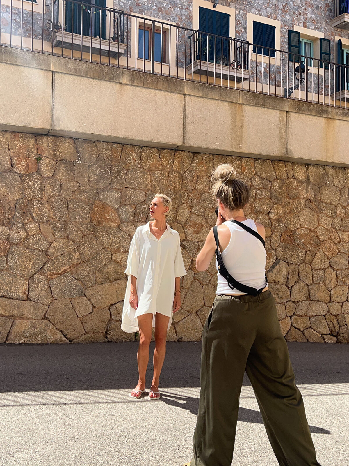 Eine Person in einem weißen Hemd steht vor einer Steinmauer, während eine andere Person sie fotografiert.