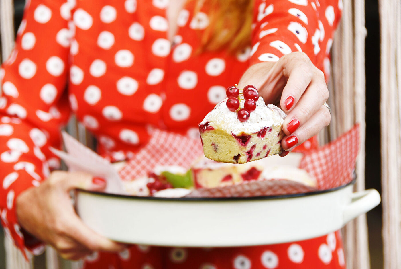 Eine Person in einem roten, gepunkteten Kleid hält ein Stück Kuchen mit Beeren in der Hand. Der Kuchen liegt auf einem weißen Teller, umgeben von roten Servietten.