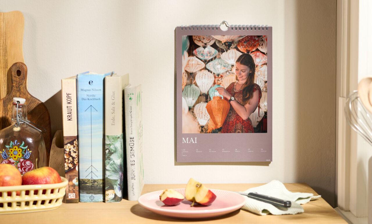 CEWE Fotokalender hängt an der Wand, zeigt den Monat Mai mit Foto einer Person vor bunten Lampions