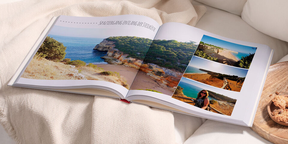Ein geöffnetes Fotobuch mit Urlaubsfotos, auf einem eleganten Tisch. Die Bilder zeigen eine Küstenlandschaft und Momente vom Strand.