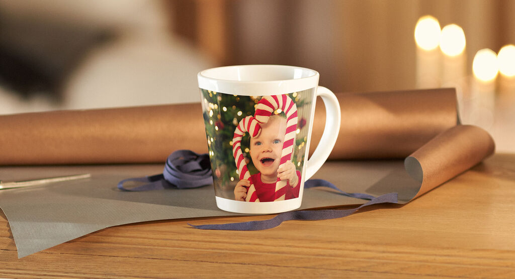 Eine festliche Tasse mit buntem Design, umgeben von Geschenkpapierröllchen und einem blauen Band auf einem Holztisch.