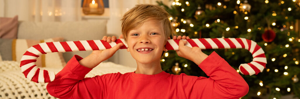 Ein Junge hält einen großen Zuckerstab hinter seinem Kopf. Im Hintergrund leuchtet ein festlich geschmückter Weihnachtsbaum.