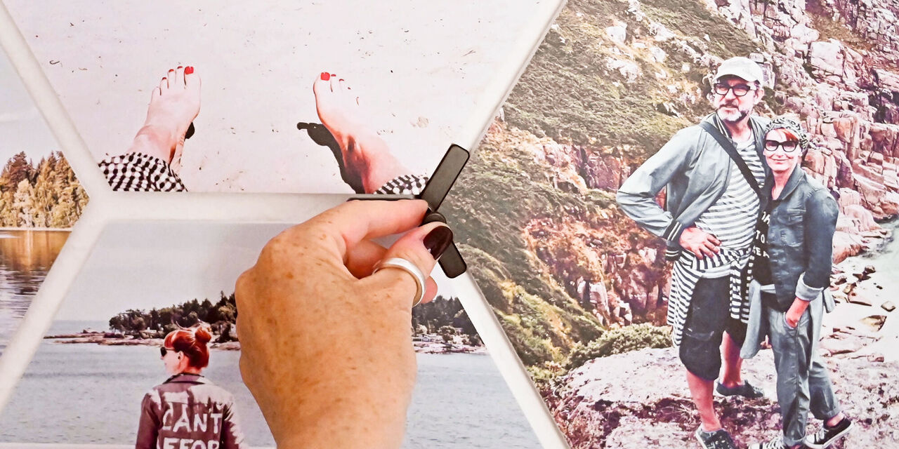 Eine Hand befestigt Fotos an einer Wand aus Holz. Die Bilder zeigen verschiedene Urlaubs- und Naturmomente.