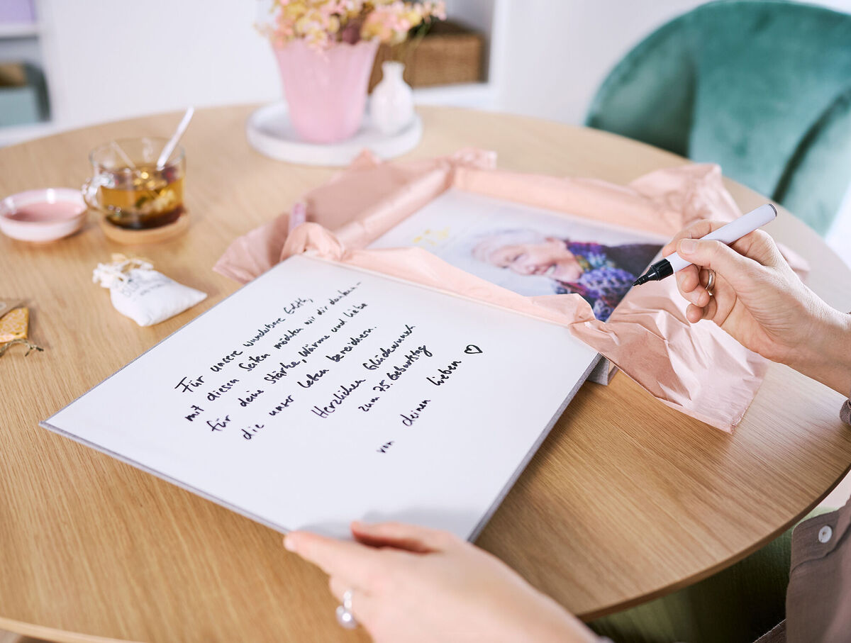 Eine Frau sitzt an einem Tisch und beschriftet den Deckel einer Geschenkbox mit persönlichen Worten.