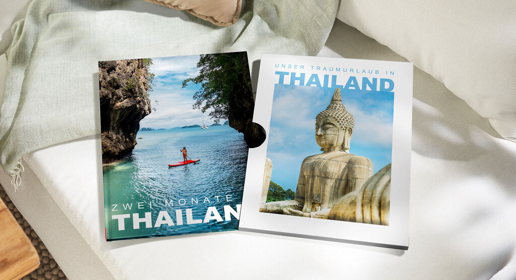 Ein CEWE FOTOBUCH Thailand mit Kajak- und Buddha-Motiv auf den Covern herausgezogen aus einem Schuber