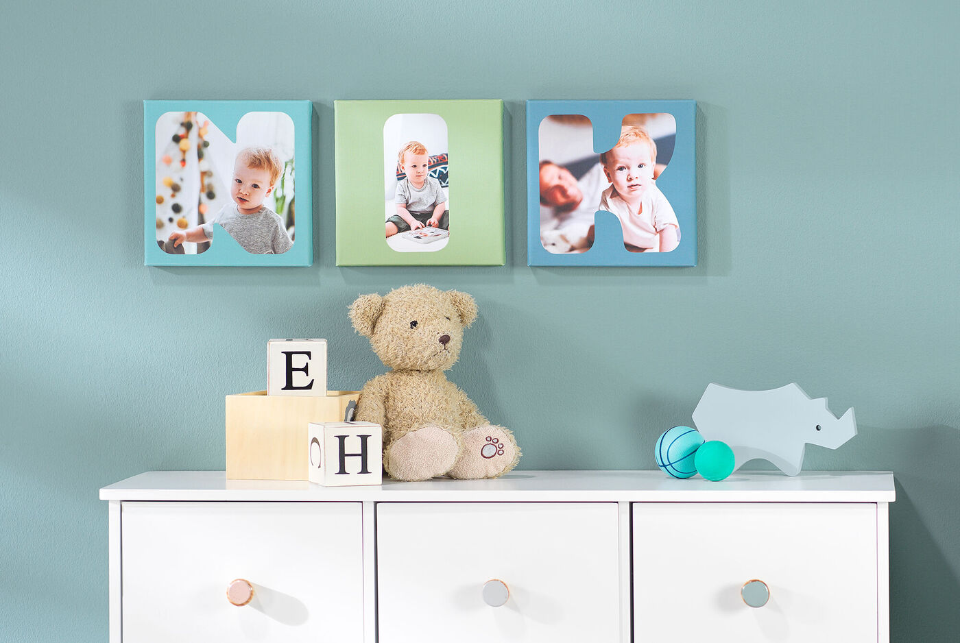 Drei Bilder in bunten Rahmen an einer Wand über einer weißen Kommode mit einem Teddybären und Spielzeug.