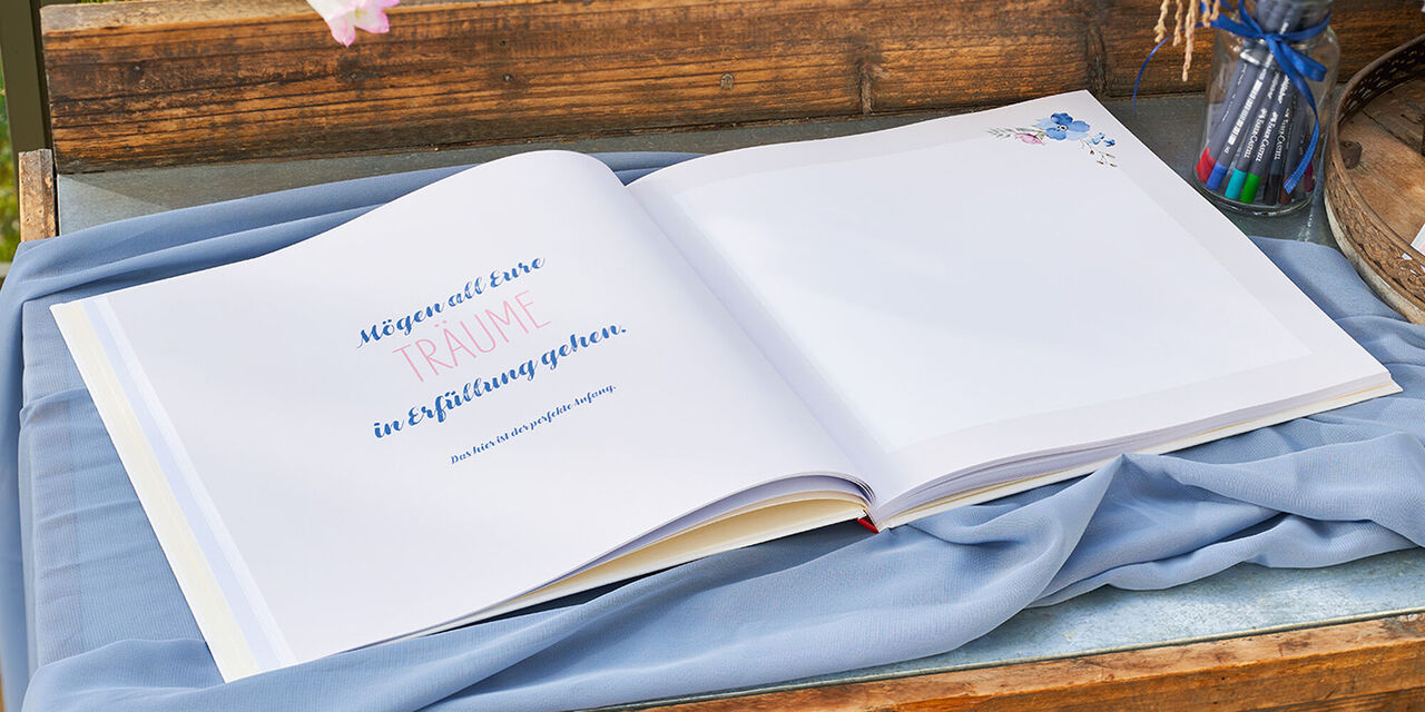Das aufgeklappte Gästebuch liegt auf einem Tisch. Auf der linken Seite steht ein klassischer Hochzeitsspruch. Die rechte Seite bietet Platz für persönliche Botschaften. Rund um das Buch befinden sich Stifte, Fotosticker und Blumendekoration.