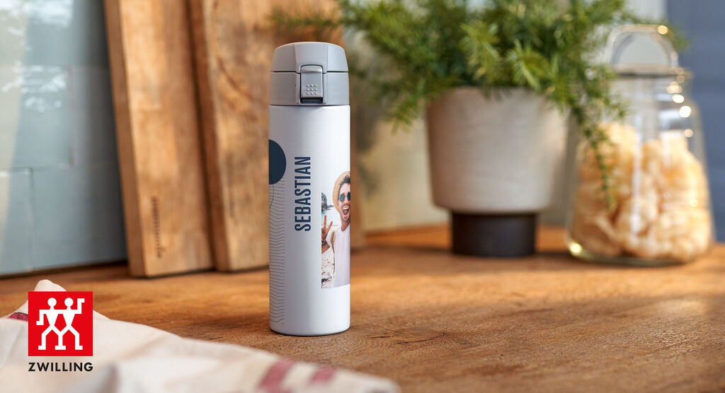 Isolierflasche mit Foto von Sebastian auf einem Holztisch, umgeben von Pflanzen und Gläsern
