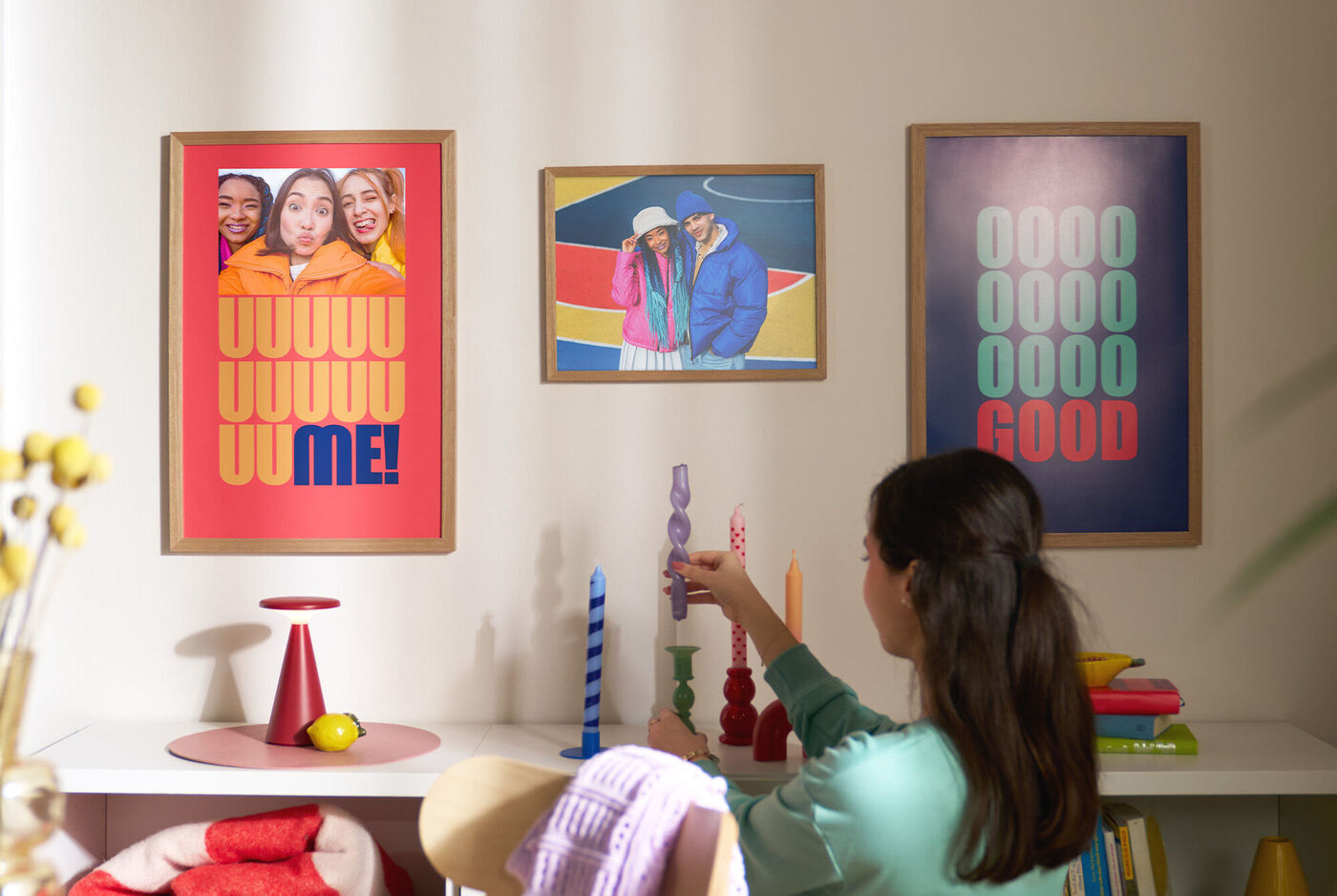 Eine Frau dekoriert ein Wohnzimmerregal. An der Wand hängen drei Poster, die mit Fotos und Schriftzügen gestaltet sind.