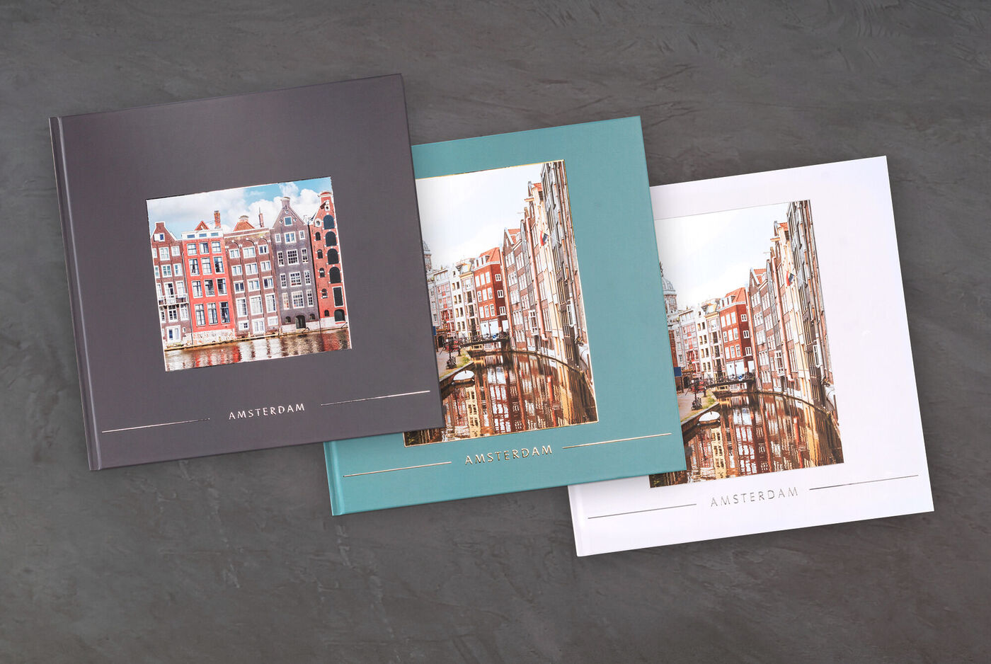Drei verschiedene CEWE Fotobücher mit Bildern von Amsterdam, jedes mit einem unterschiedlichen Farbschema.