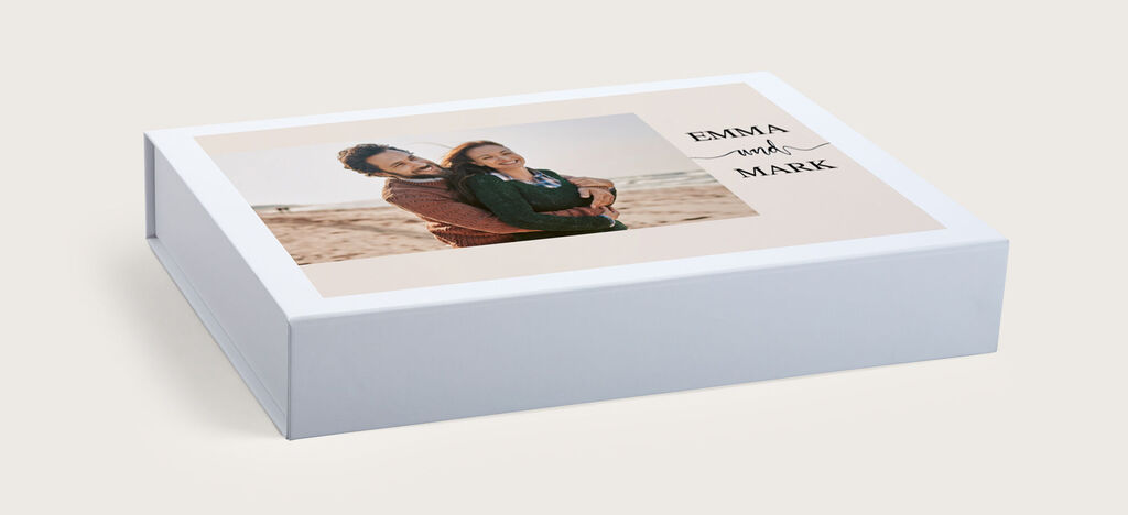 Wundervolles CEWE Fotobuch mit einem glücklichen Paar am Strand. Es zeigt die Namen EMMA und MARK auf dem Cover, umrahmt von einem sanften Design.