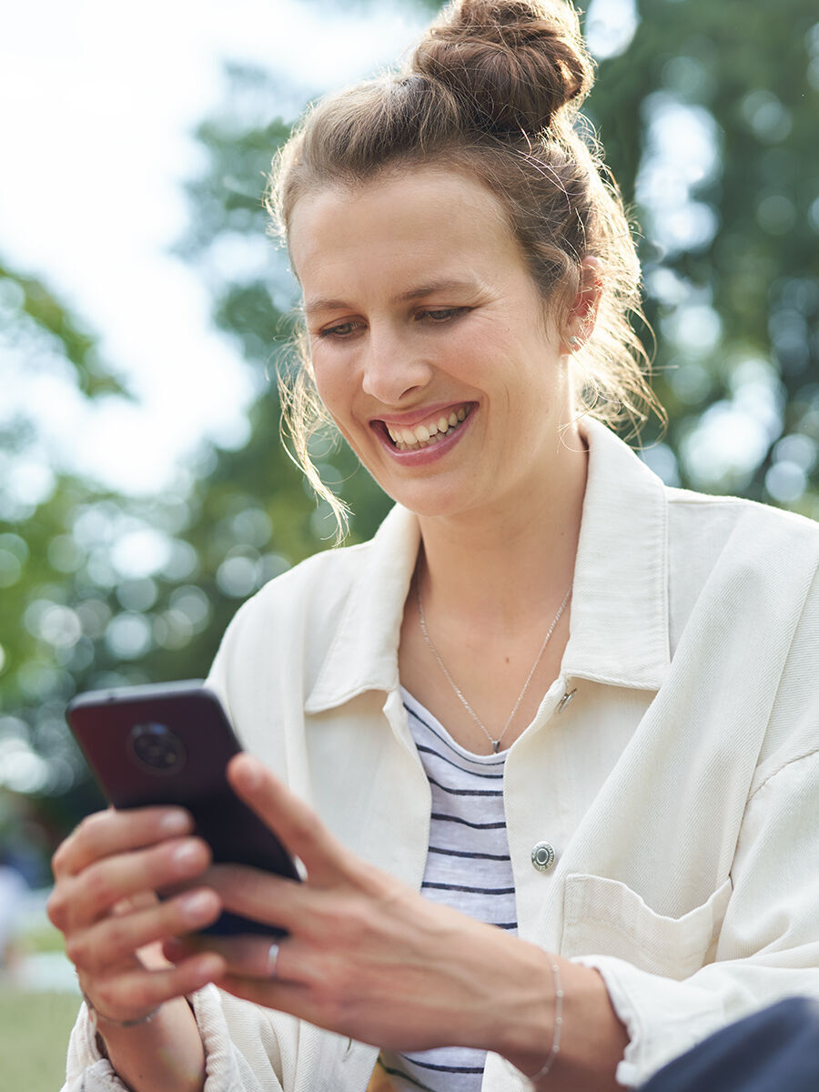 Eine Person sitzt im Freien und schaut auf ihr Smartphone. Sie trägt eine helle Jacke und ein gestreiftes Shirt. Die Umgebung ist grün und sonnig.