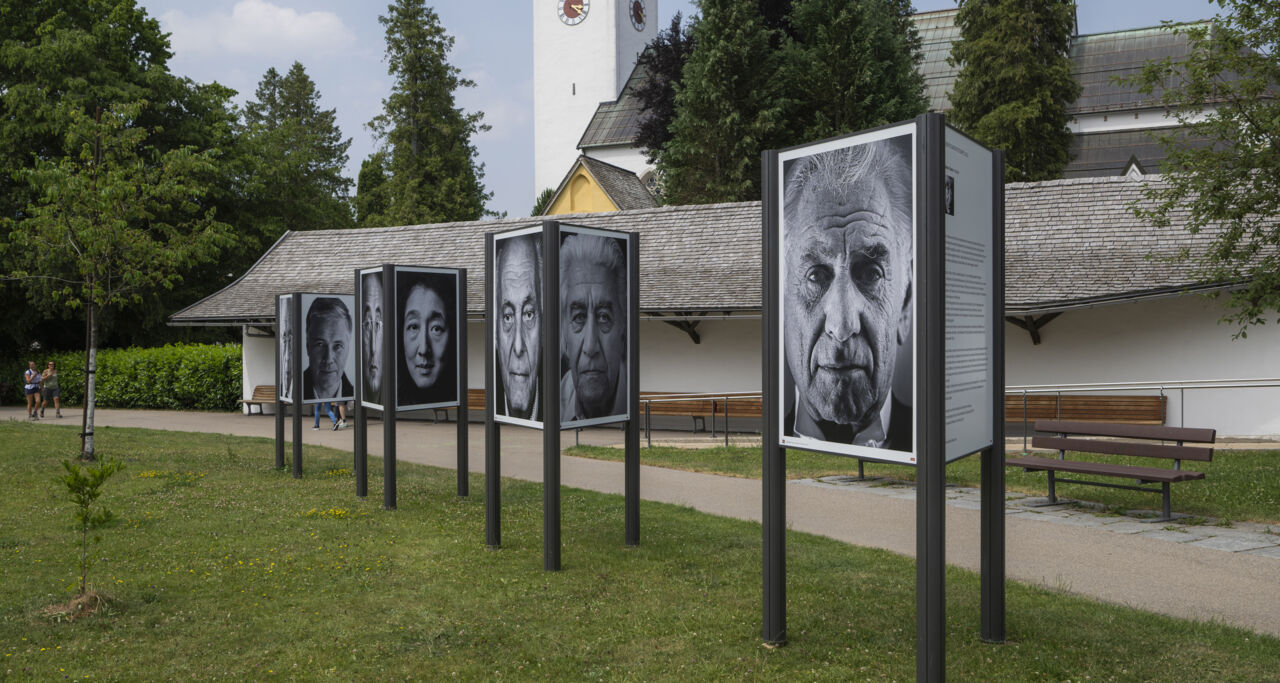 Drei große Aufsteller auf einer Wiese zeigen verschiedene Schwarz-Weiß-Porträts. Im Hintergrund ist ein Kirchturm.